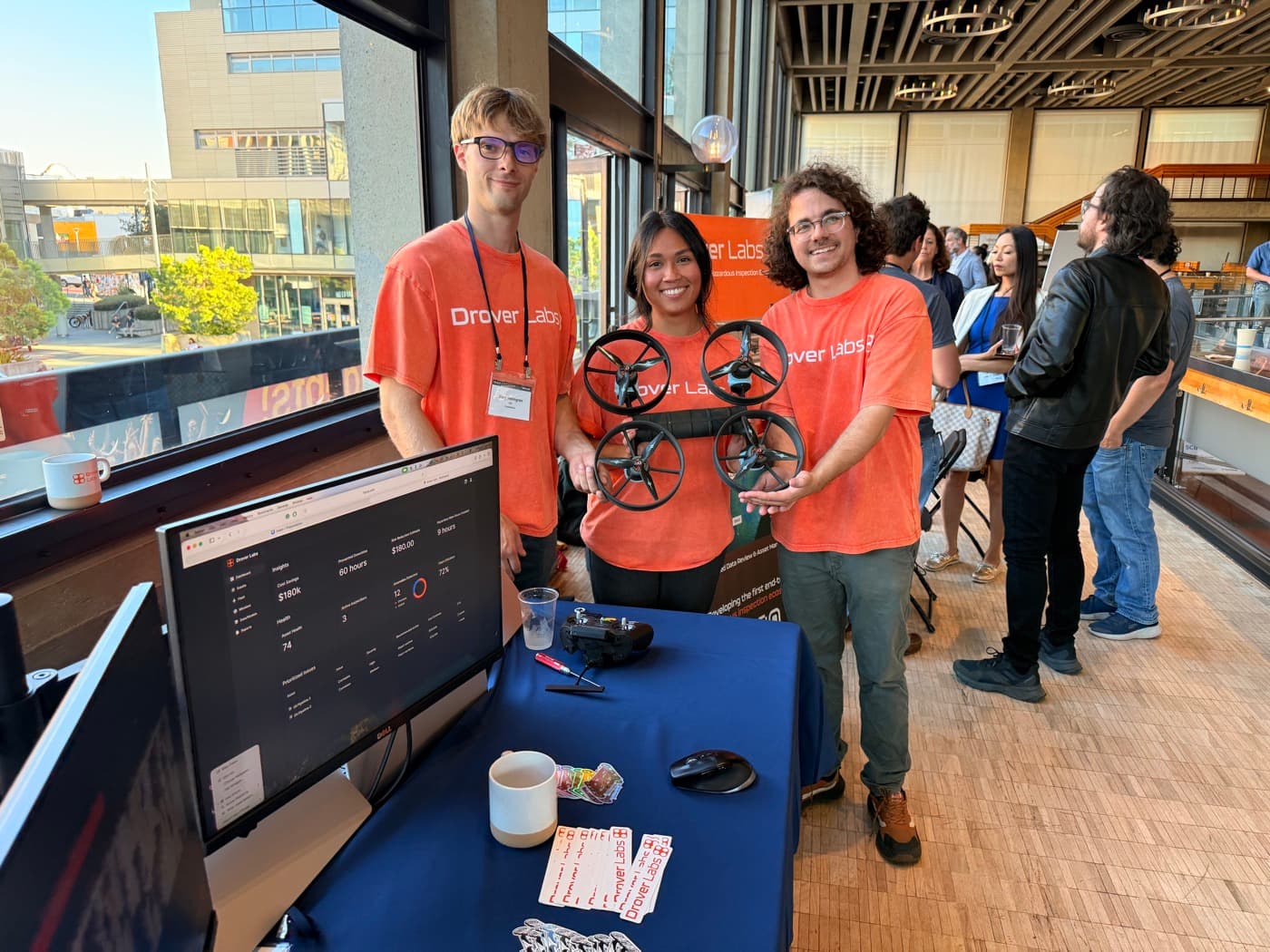 The three Drover Labs founders in orange shirts holding an early robot prototype at a demonstration booth